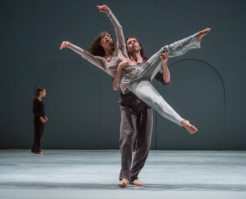 A man holds a woman off kilter. One hand supports around her waist while another hand holds her thigh. Her long brown hair covers her face.